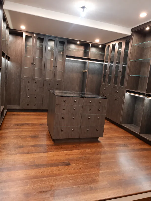 Dark wood-grain dressing room with glass cabinet doors, glass shelves, lighting and a glass-top drawer island