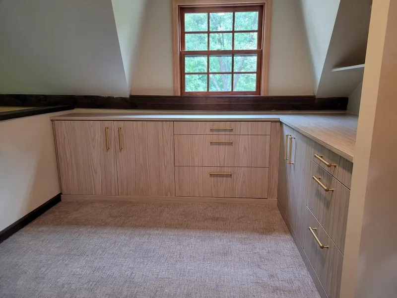 Light wood-grain attic cabinetry with paneled doors, drawers and a countertop fitted beneath a window and sloping walls