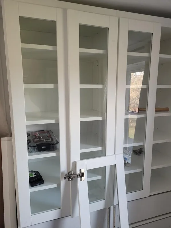 During installation: white glass-front storage cabinet with tall adjustable shelves and lower drawers while the doors are being fitted