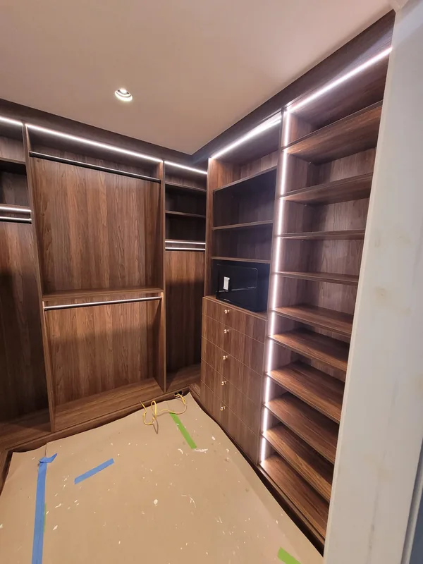 Near completion: wood-grain walk-in closet with continuous lighting, shoe shelves and hanging rails, with protective floor covering still in place