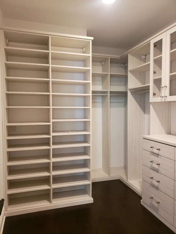 White walk-in closet with a tall shoe tower, wood-accented cabinetry and dark flooring