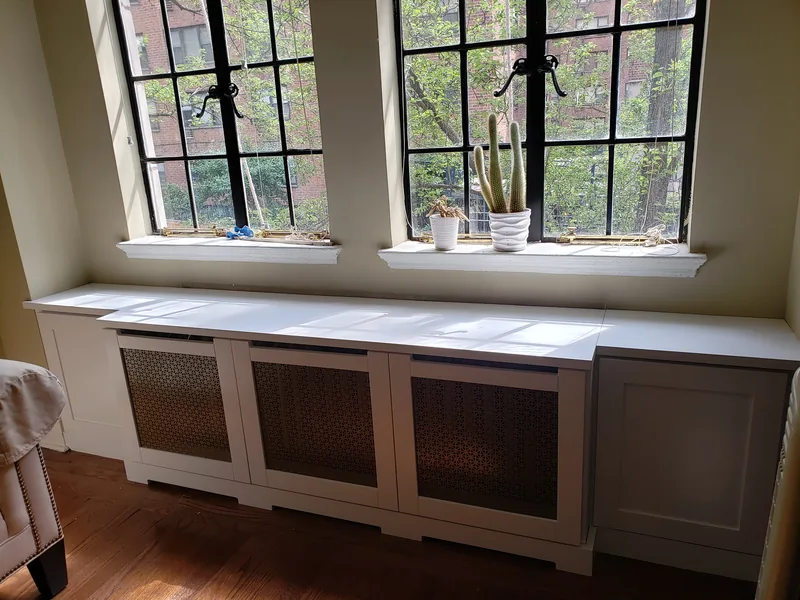 Long white radiator cover with three decorative grille panels and side cupboards below two windows